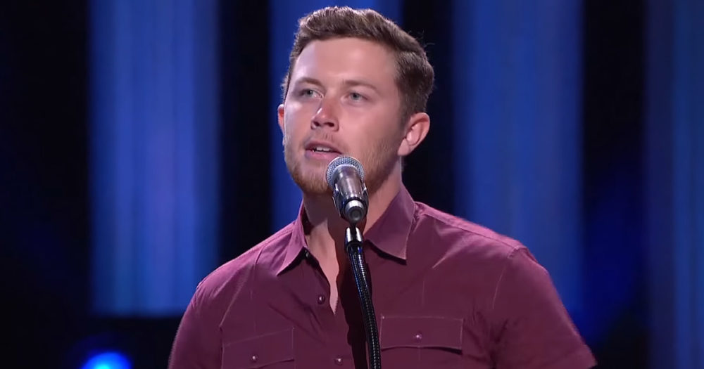 A man in a maroon shirt sings into a microphone on stage with blue lighting.